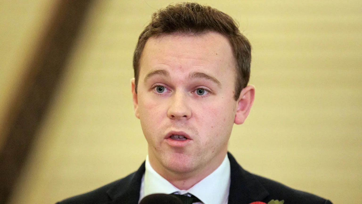  Eoin Tennyson speaking. He has brown hair and is wearing a black blazer with white shirt and black tie.