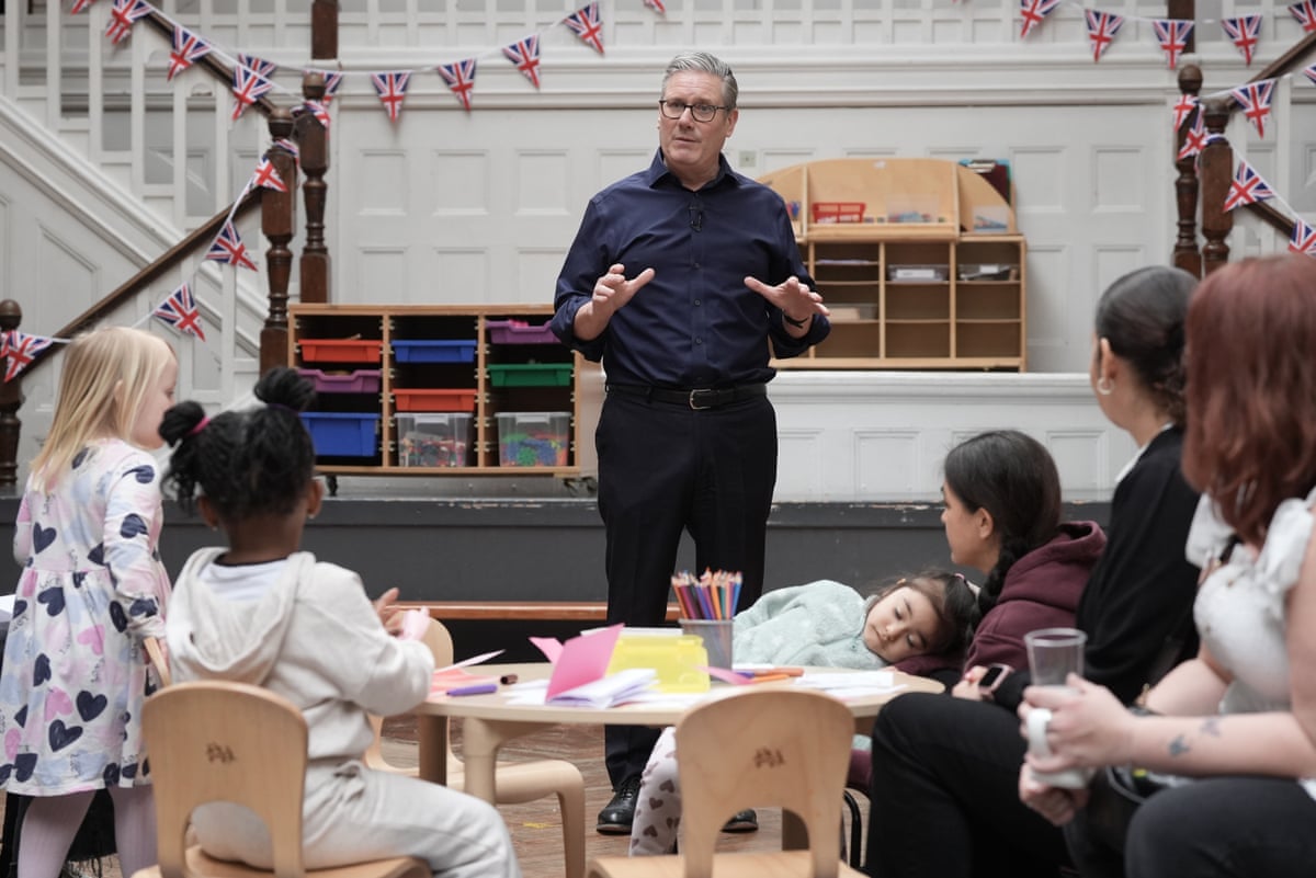 Keir Starmer delivering a speech at a school with small children sat on a table in front of him. One of the children is slumped to one side asleep. 