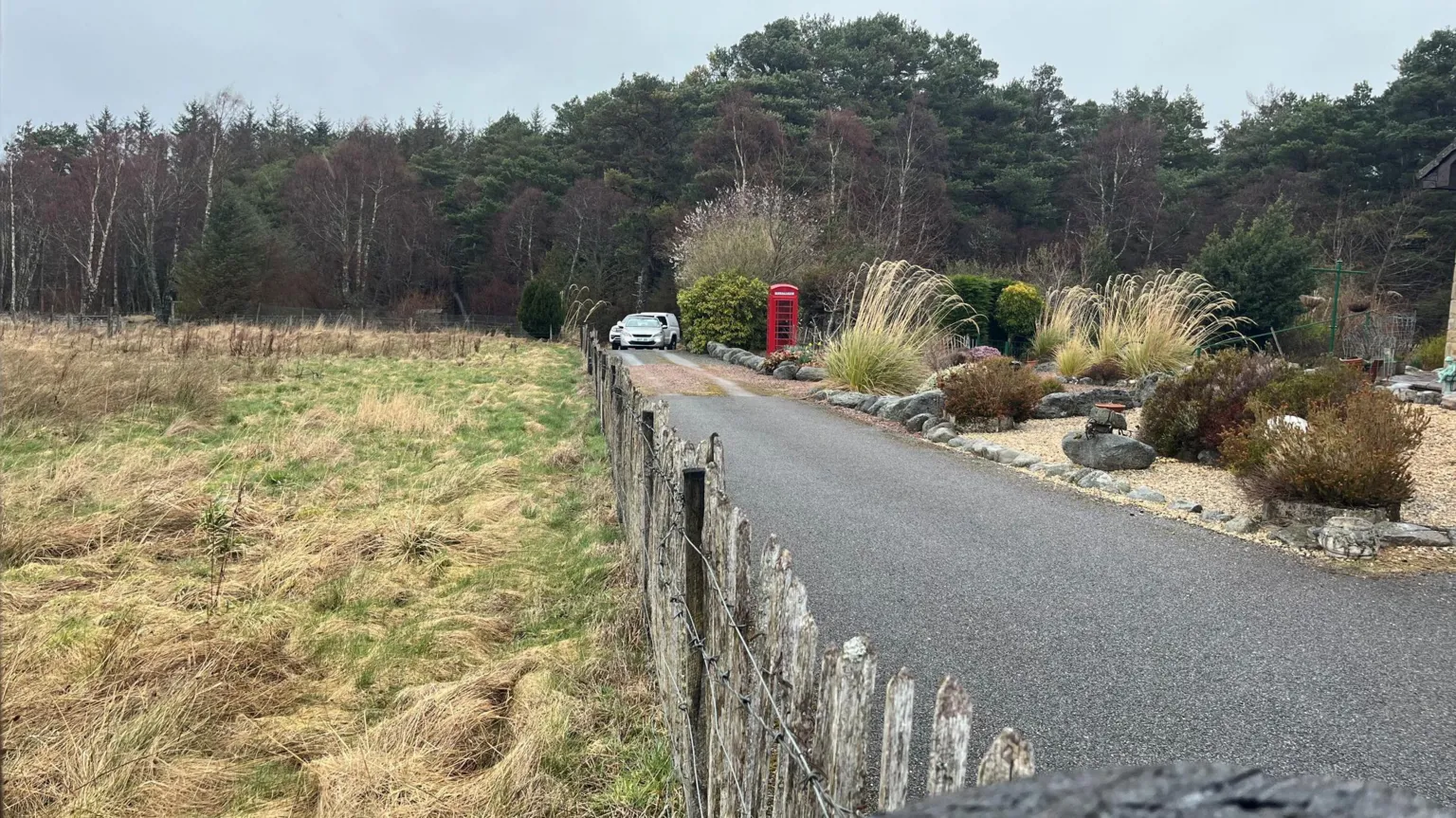 A rural road with a field on one side and a large garden on the other. At the end of the road are parked two vehicles and there is a large area of woodland.