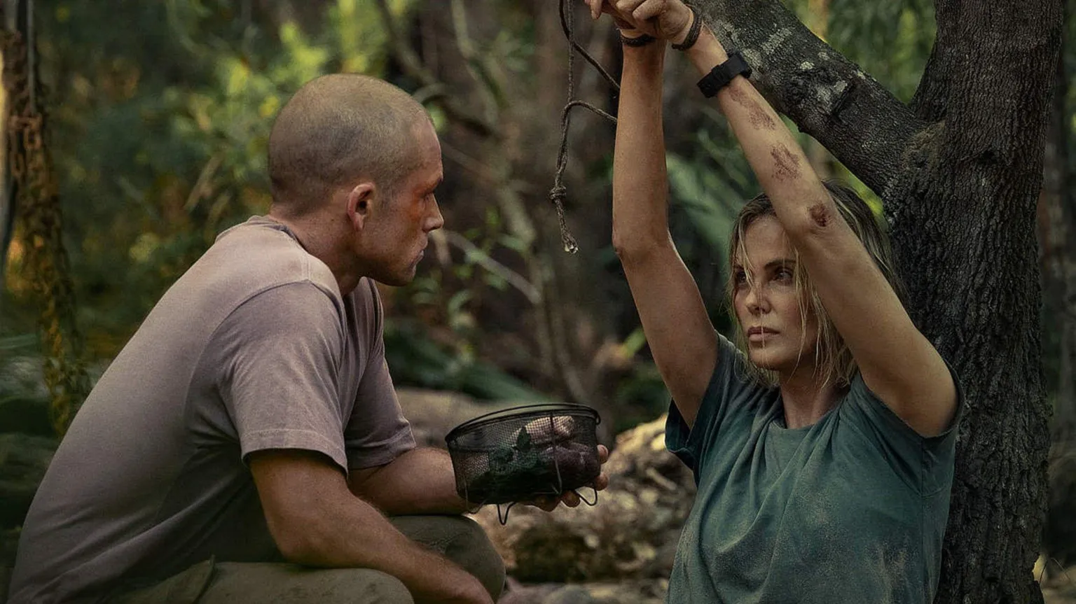 KANE SKENNAR/NETFLIX Egerton and Theron. Theron's arms are tied above her head and attached to a tree branch. She wears a blue t-shirt. Egerton wears a grey/purple t-shirt