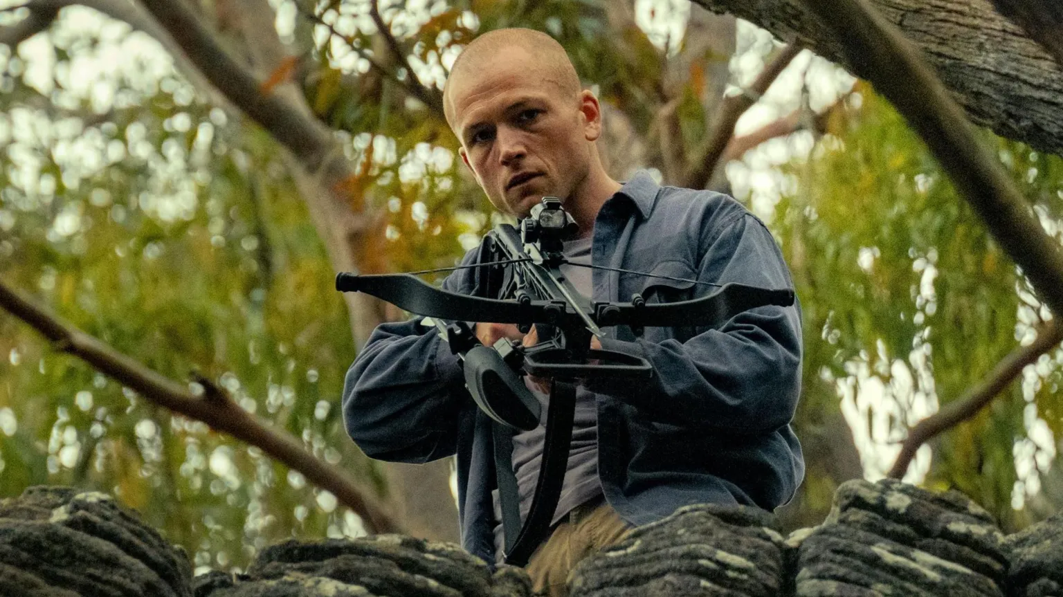 KANE SKENNAR/NETFLIX Egerton is above the camera in a forest, aiming a crossbow. He wears a blur shirt over his grey t-shirt