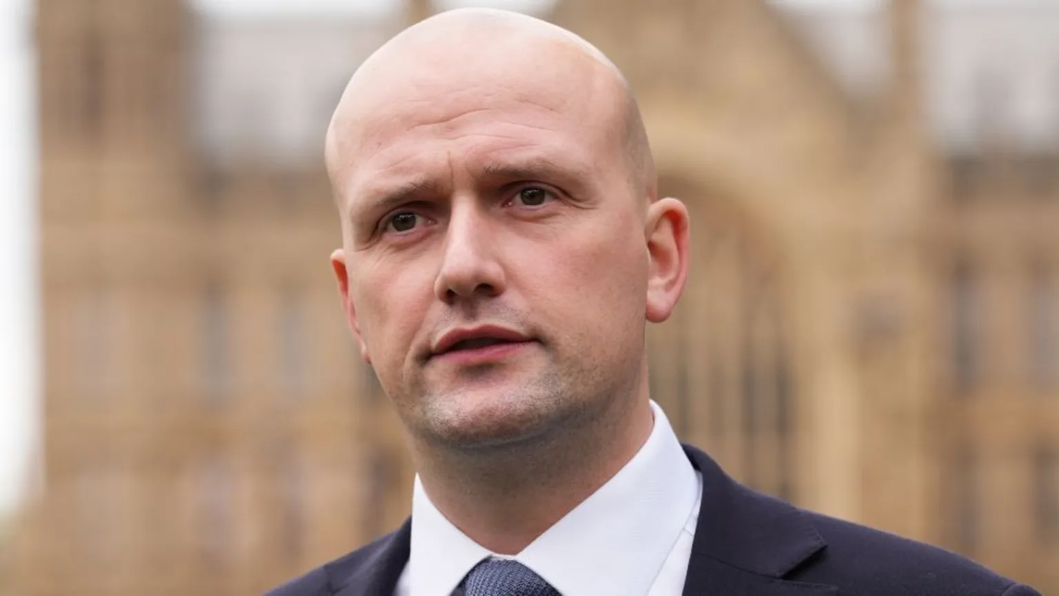 Stephen Flynn, who is bald, outside the UK Parliament, a large stone building out of focus in the background. He is wearing a dark suit, white shirt and blue tie.