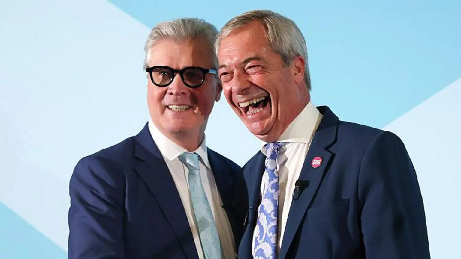 Malcolm Offord and Nigel Farage standing close together on a stage. They both have short, grey hair and are wearing blue suits and cream or white shirts. Offord has dark-rimmed glasses and is wearing a light blue tie. He is smiling while Farage, who is laughing, is wearing a paisley-patterned blue and white tie.
