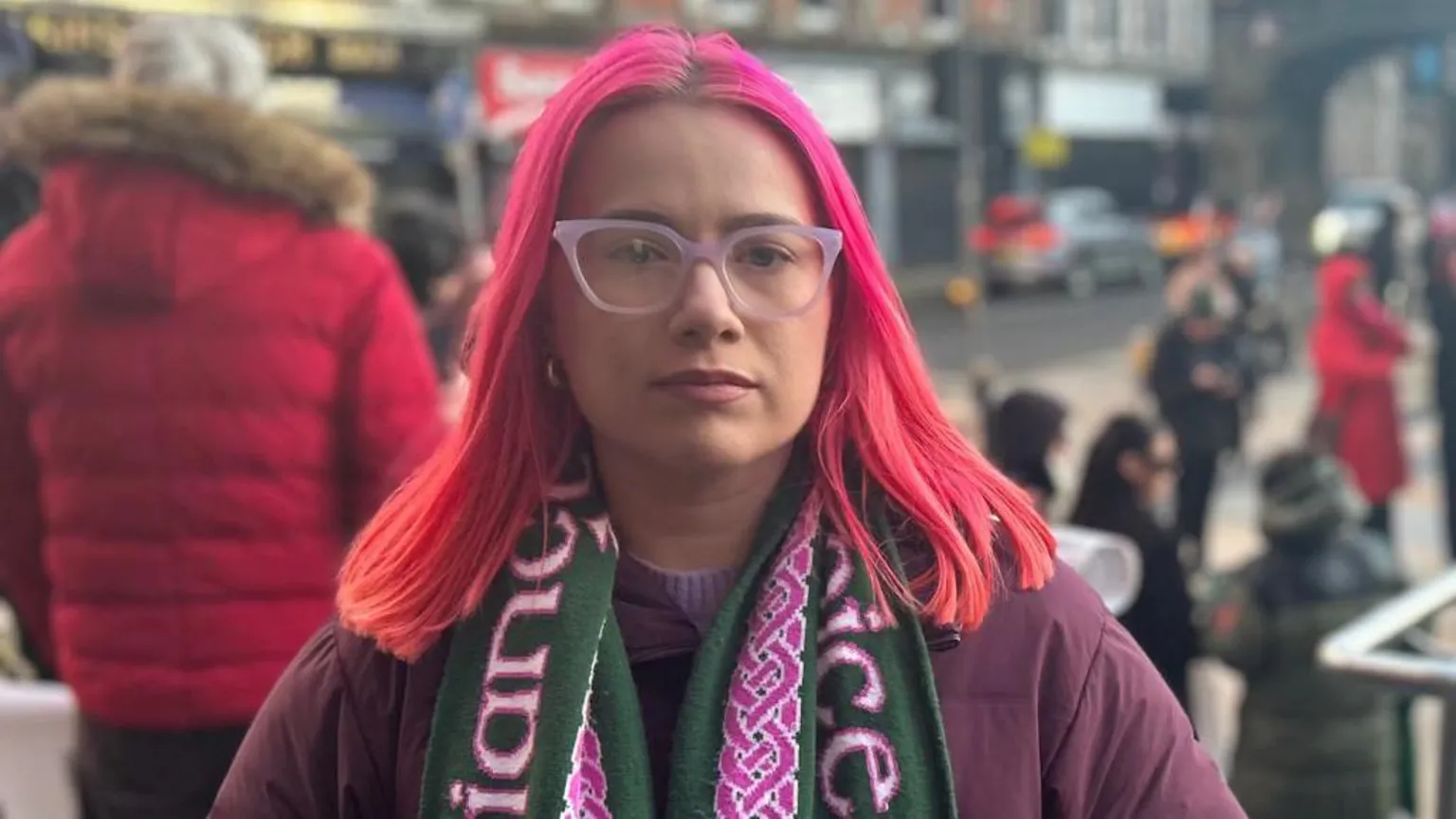 A woman with pink hair is wearing a pink pair of glasses, a purple jacket and a green and purple scarf. She is standing on steps in front of a small crowd of people