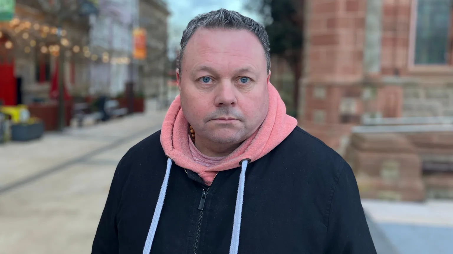 a man with short hair stands in Derry's Guildhall Square. Some lit up buildings are in the background to his right, while on left is the red bricks of the Guildhall. He is wearing a black jacket over a pink hoodie