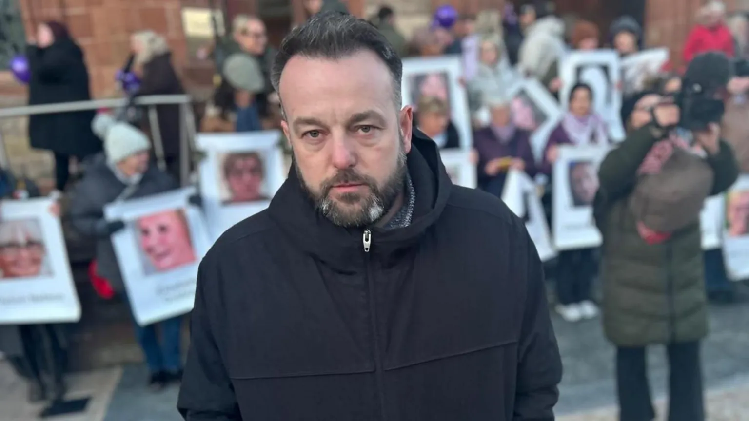Foyle SDLP MP Colum Eastwood stands in front of Derry's Guildhall. He is wearing a black coat, behind him are a number of people holding posters