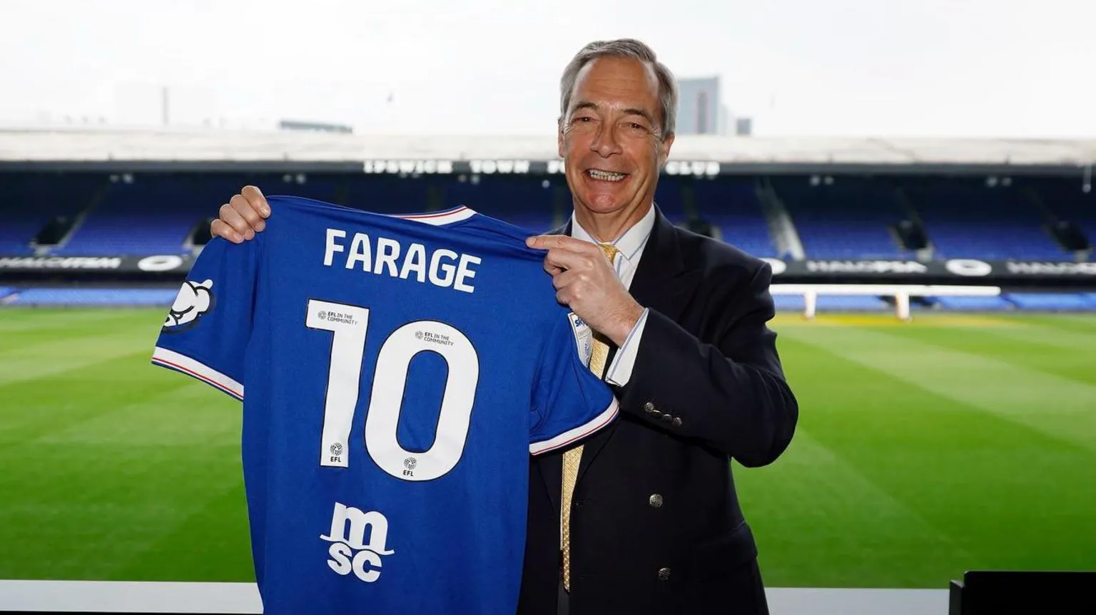 Reform Nigel Farage poses with a blue Ipswich Town home shirt with 'FARAGE 10' on the back. He is standing next to the pitch at the Portman Road stadium.