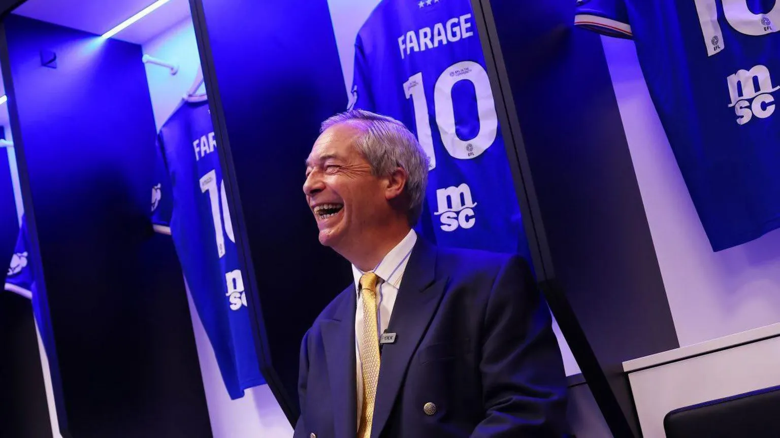 Reform UK Nigel Farage in a navy suit and a yellow tie sits in Ipswich Town's home dressing room surrounded by personalised 'FARAGE 10' Ipswich shirts. He is smiling and laughing as the picture is taken.