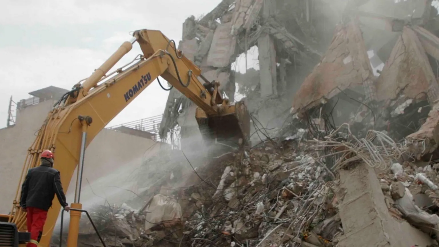 The shattered concrete and twisted metal remains of a building. A man in a black jacket with orange trousers and hard hat operates a digger to move rubble.
