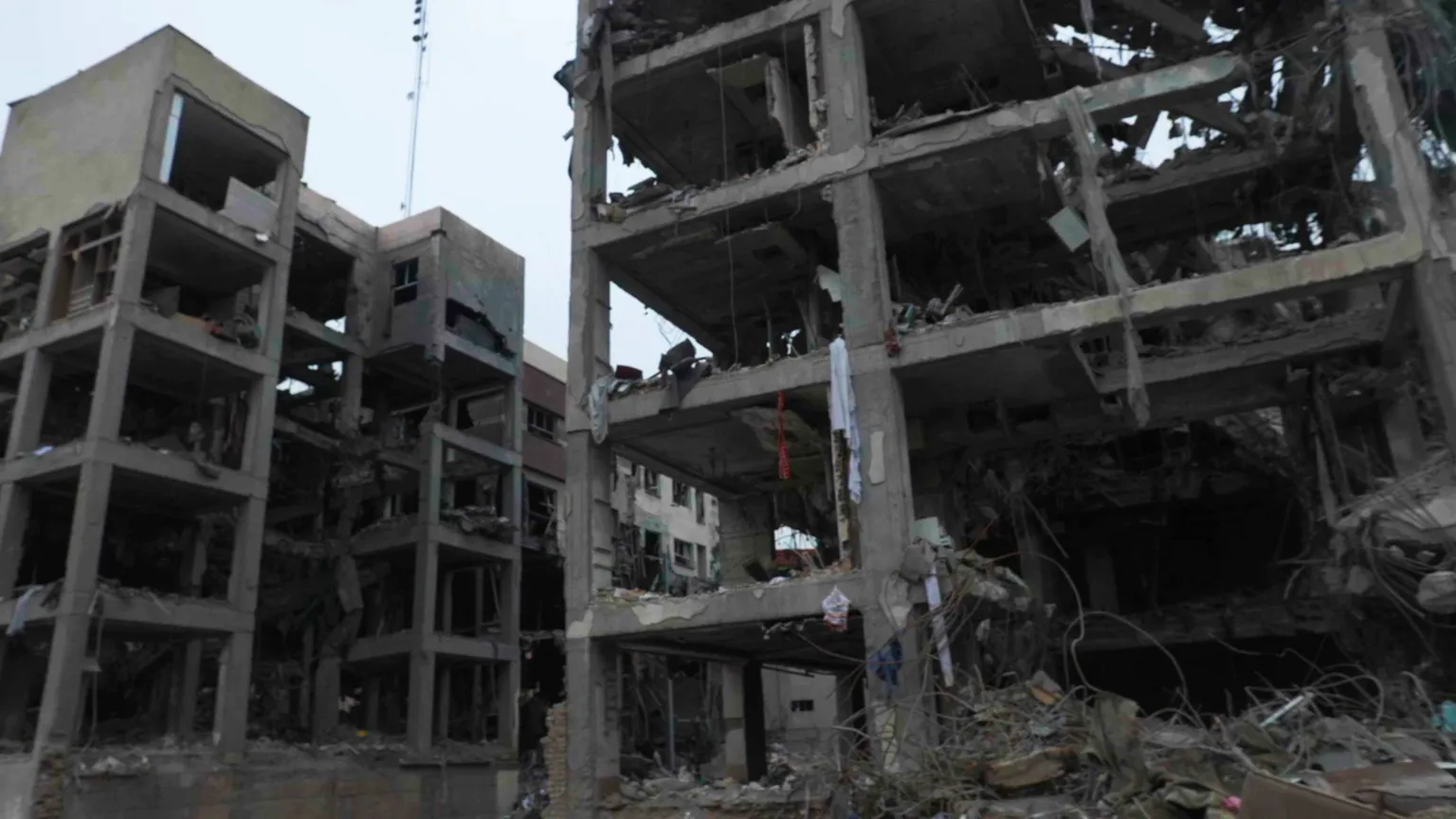 Two destroyed buildings, four to five stories high, with the windows and walls blown out. Some concrete pillars and floors remain. There are a few bits of fabric hanging off the structure and some twisted metal on the ground.