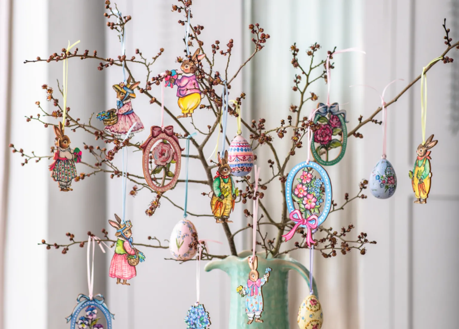 Amy Swann A tree cutting with small buds on it in a green vase with Easter decorations made by Amy hanging from the branches. The decorations are woodcuts of anthropomorphised rabbits in colourful clothing, eggs, and bunches of flowers. They are all colourful with pink, light blues, yellow and light green featuring in the designs.