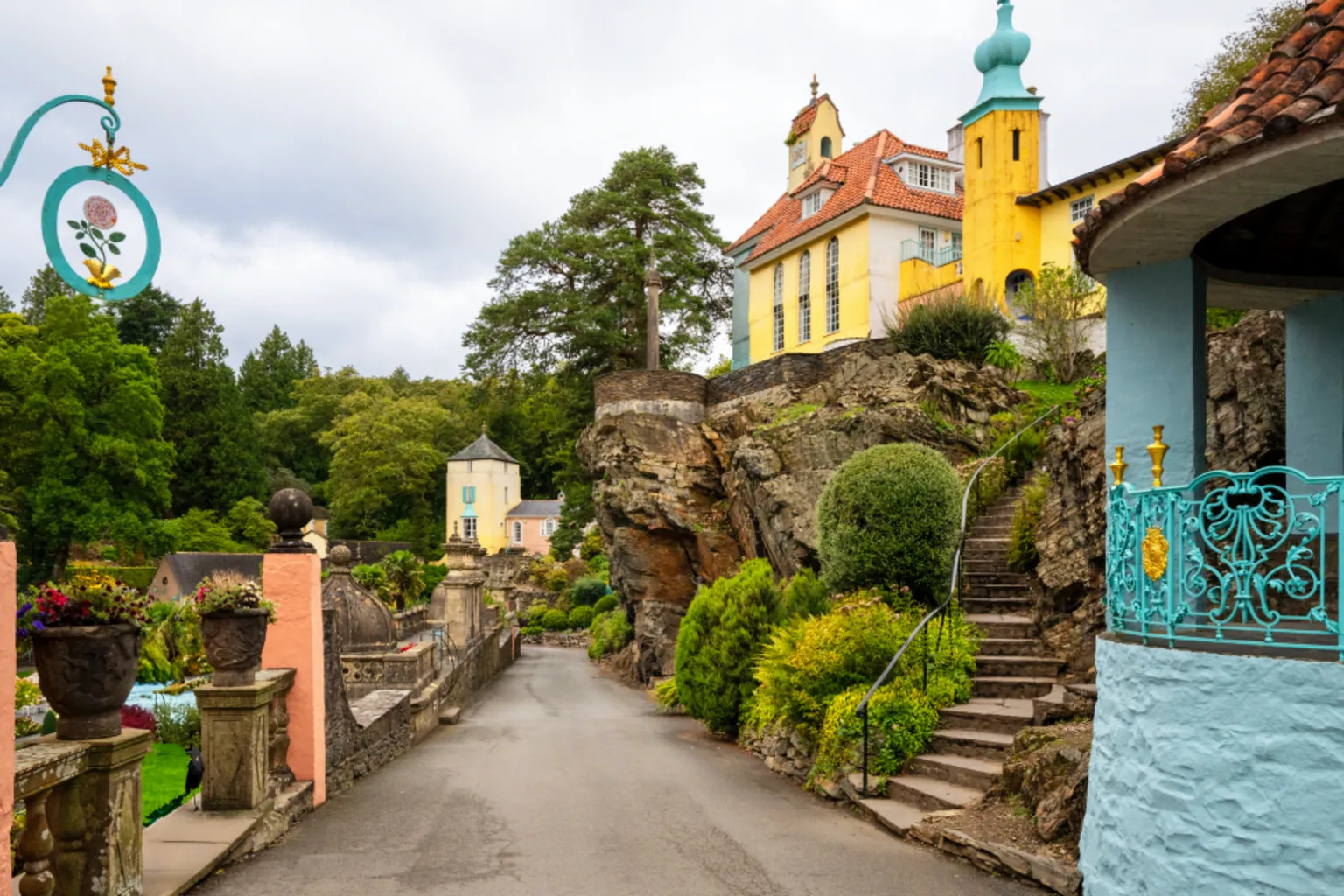  Buildings in Portmeirion. A road runs in the middle of the photo. To the right is a turquoise mini pavillion. Steps lead up to a yellow house with a turquoise turret and terracotta tiled roof in the Italian style. It stands on an outcrop of rock. To the left are the walls of gardens including a couple of peach painted pedestal supports. A couple of buildings painted yellow and light orange are in the distance.