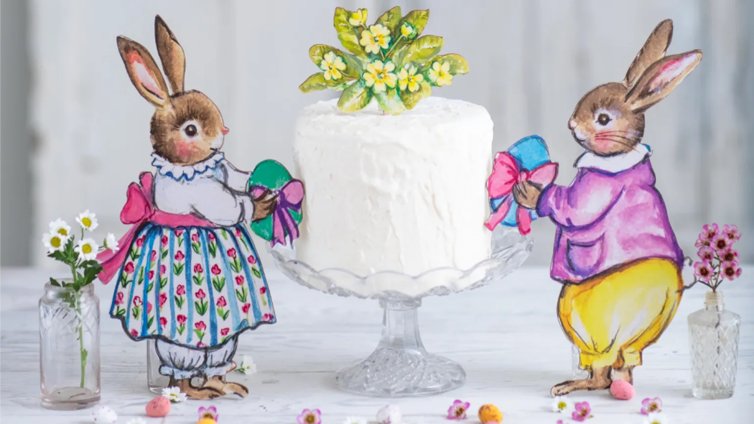 Amy Swann A white iced cake with a drawn decoration of yellow primroses with green leaves sits on top of it. It rests on a glass stand. To either side are stylised drawings of rabbits wearing colourful but old-fashioned clothes each holding a coloured egg with a bow wrapped around it which leans onto the cake. There is a small glass vase of tiny fresh flowers behind each rabbit, and some daisies and other small flowers scattered on the white tablecloth