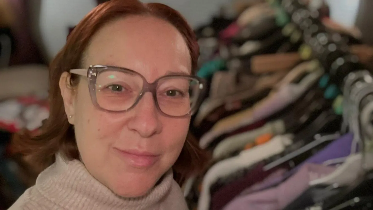 Rachel Grady Selfie photograph of Rachel Grady. She has a ginger short bob, wears square glasses, has blue eyes and smiles at the camera. The background is blurred but shows railings of clothes. 