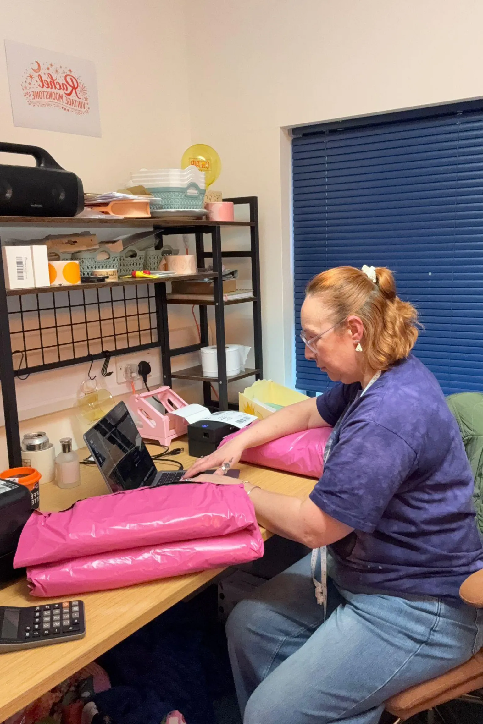 Rachel Grady Rachel Grady sits at a laptop in front of a wooden desk. Two pink parcels can be seen on the desk, and she leans her right arm on another pink parcel. She has ginger short hair, which is half pulled up into a bobble and she looks down at the laptop. Blue shutters can be seen behind her. 