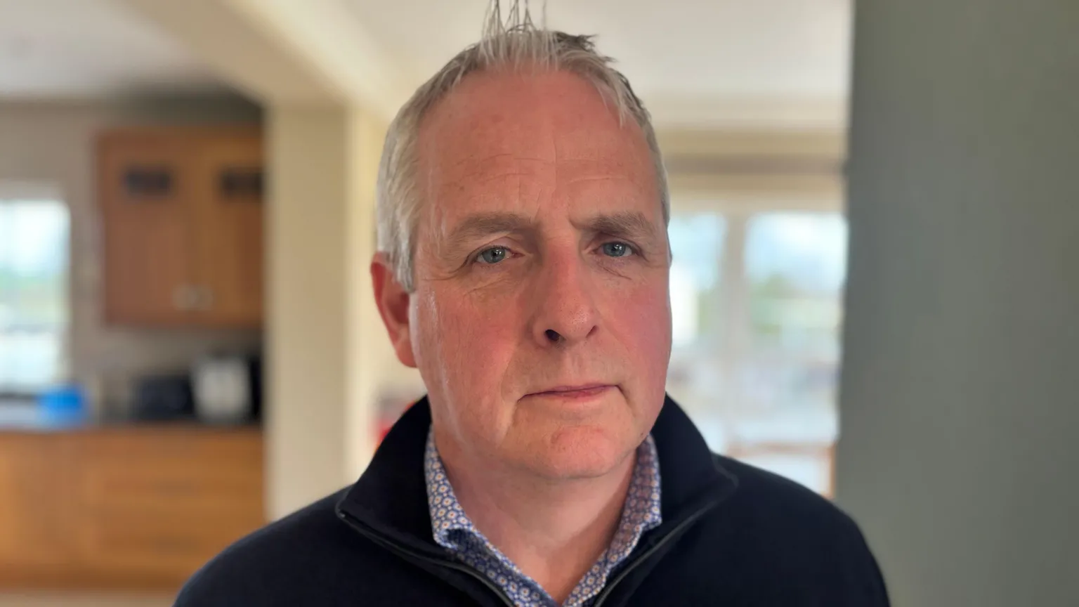A man stands in a kitchen. He has grey hair and blue eyes. He is smartly dressed in a shirt and a zip up navy jumper. His expression is somber. Behind him we can see some wooden kitchen cabinets and a pale green wall. 
