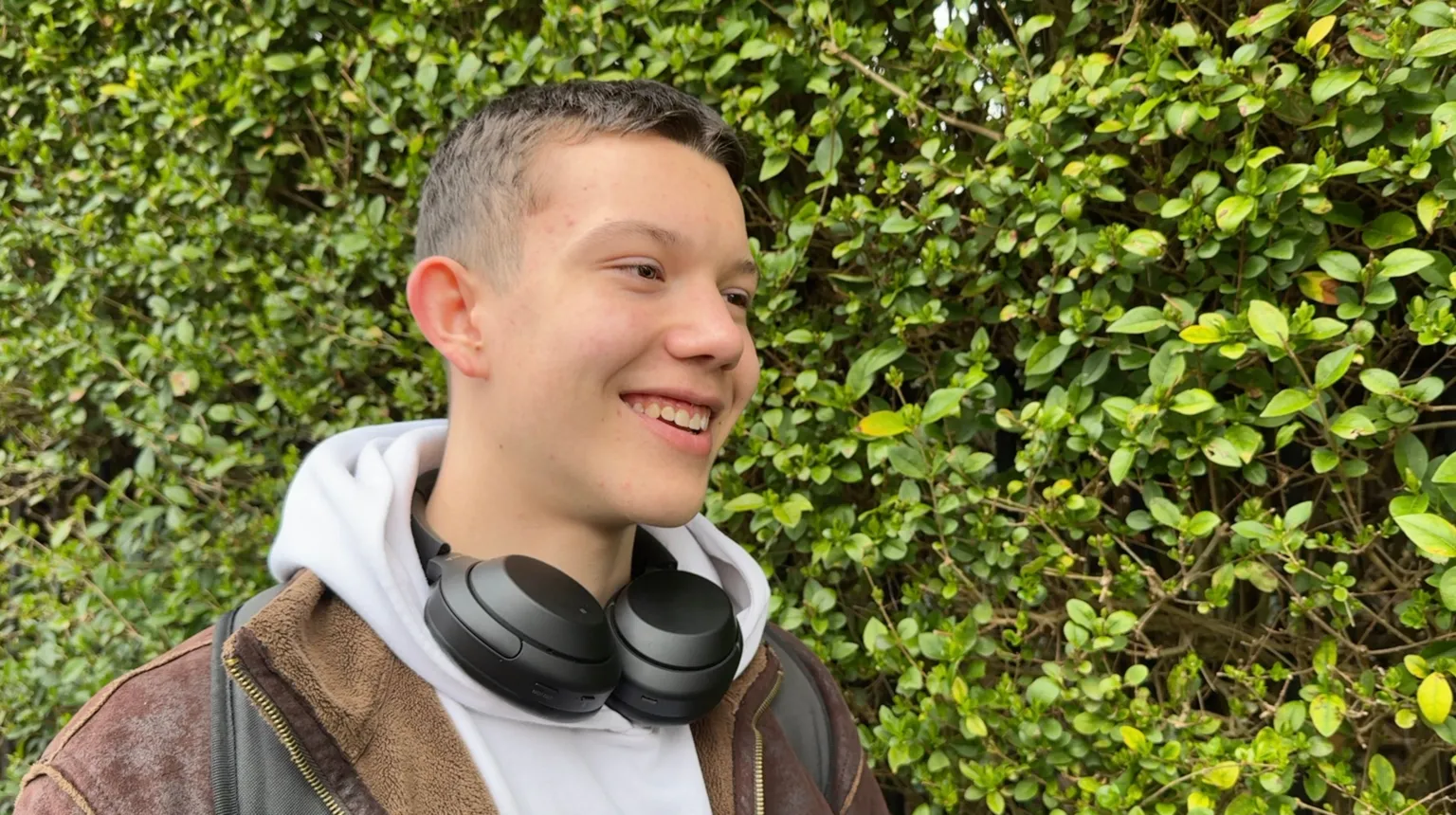 Image shows a student called Mavi. He is looking off to the left side. He has brown short hair. He is smiling. He had a white hoodie on and a brown jacket over the top. You can see the straps of his backpack which are black. He has a pair of black headphones round his neck. The whole background is filled with a large green bush. 