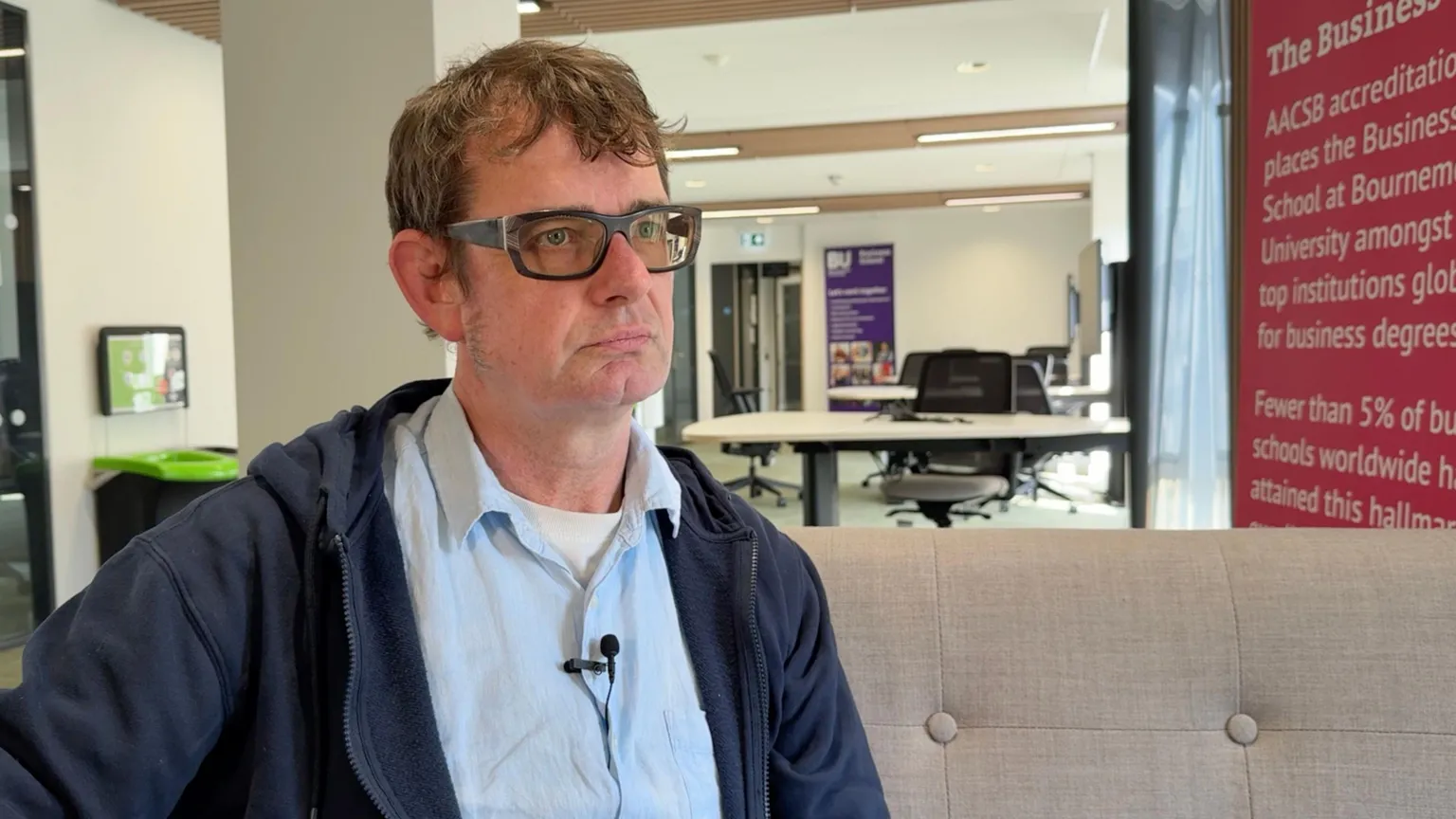 The image shows a Senior Lecturer in Economics at the University of Bournemouth. He is sat on a beige sofa. Behind him are two white desks with black office chairs. To the right of him is a pink banner. He has short brown hair and black glasses. He is wearing a blue shirt with a white top underneath. Over the top, he has a blue hoodie. 