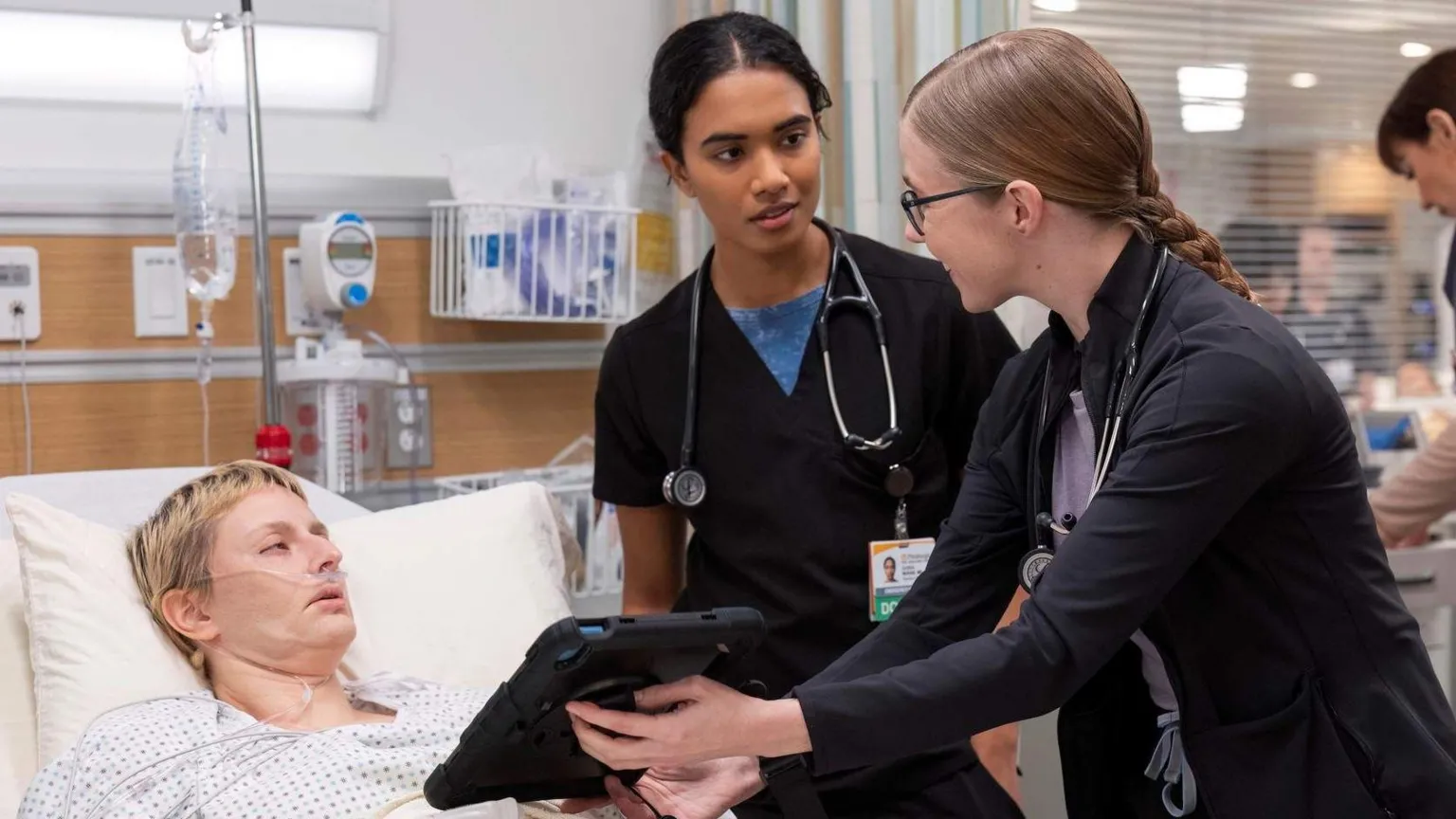 HBO Max Unknown actor lying in hospital bed with Dr Samira (Supriya Ganesh, centre) and Dr Mel (Taylor Dearden, right) at her bedside showing her a screen on a tablet