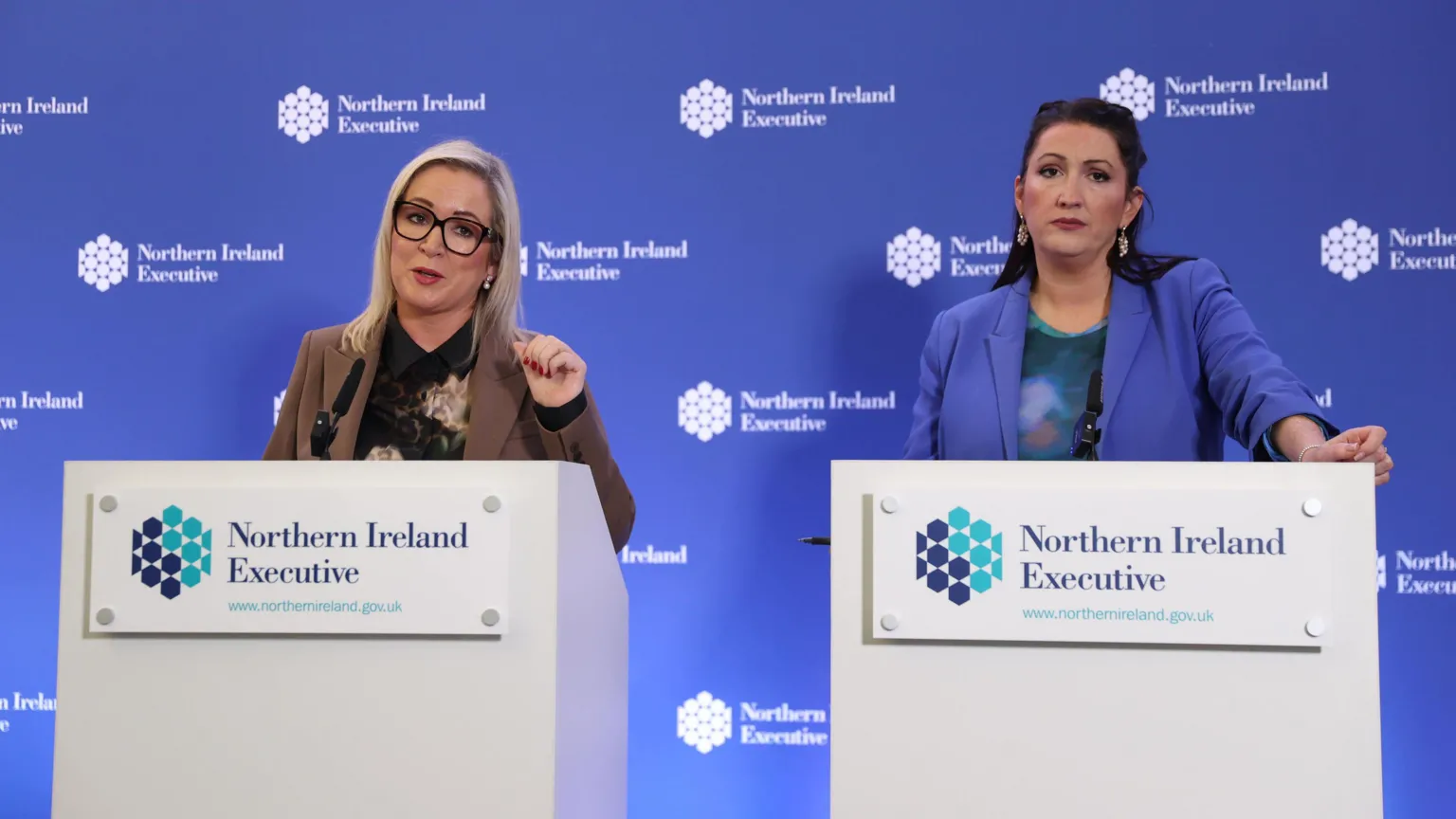  A picture of Michelle O'Neill, a blonde haired woman with glasses and wearing a brown blazer, and Emma Little-Pengelly, a black haired woman wearing a blue blazer, standing beside each other at podiums at a press conference in front of a blue background.
