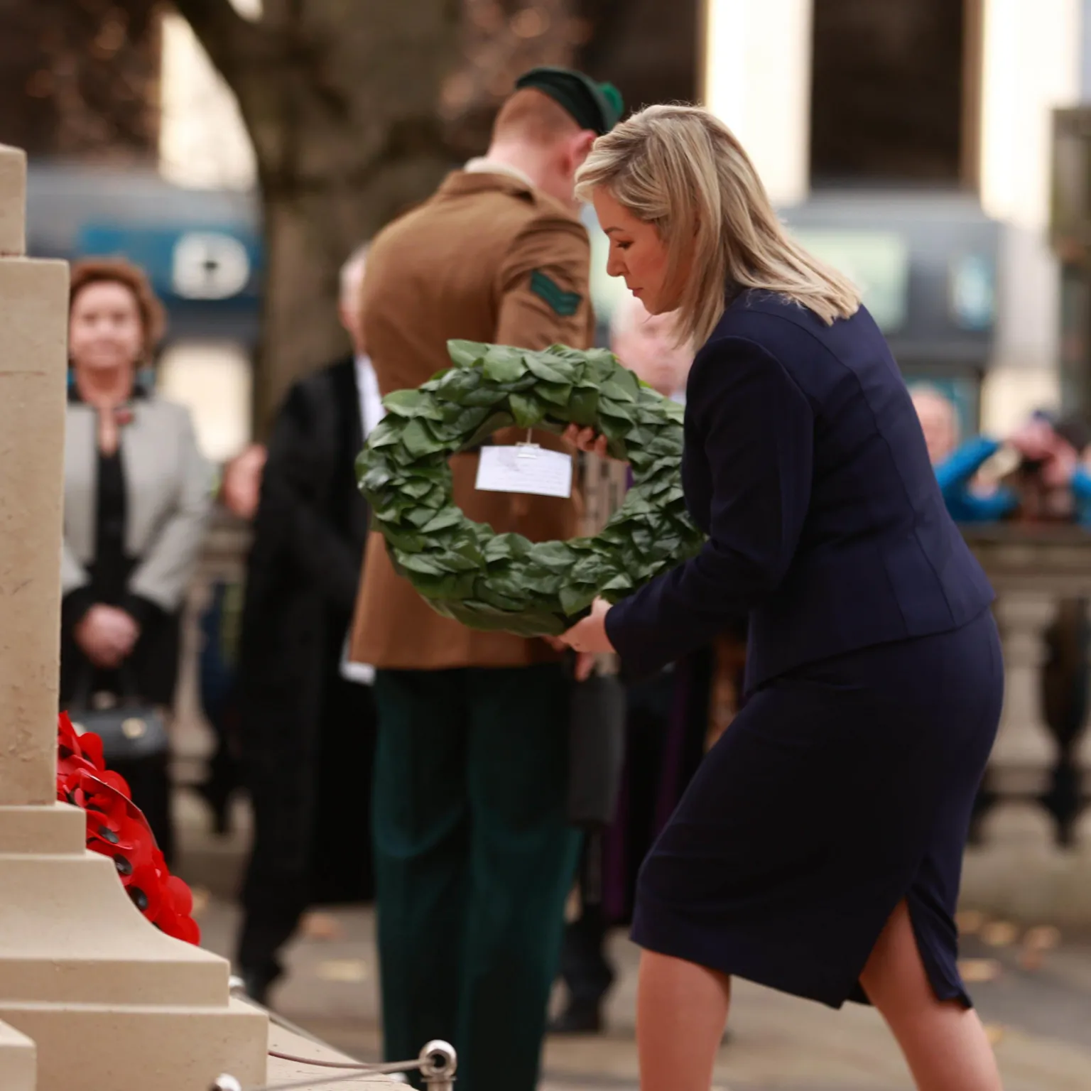  Michelle O'Neill, a woman in a dark suit, is placing a green wreath at a stone memorial. A soldier stands behind her, and a small crowd is watching quietly in the background.