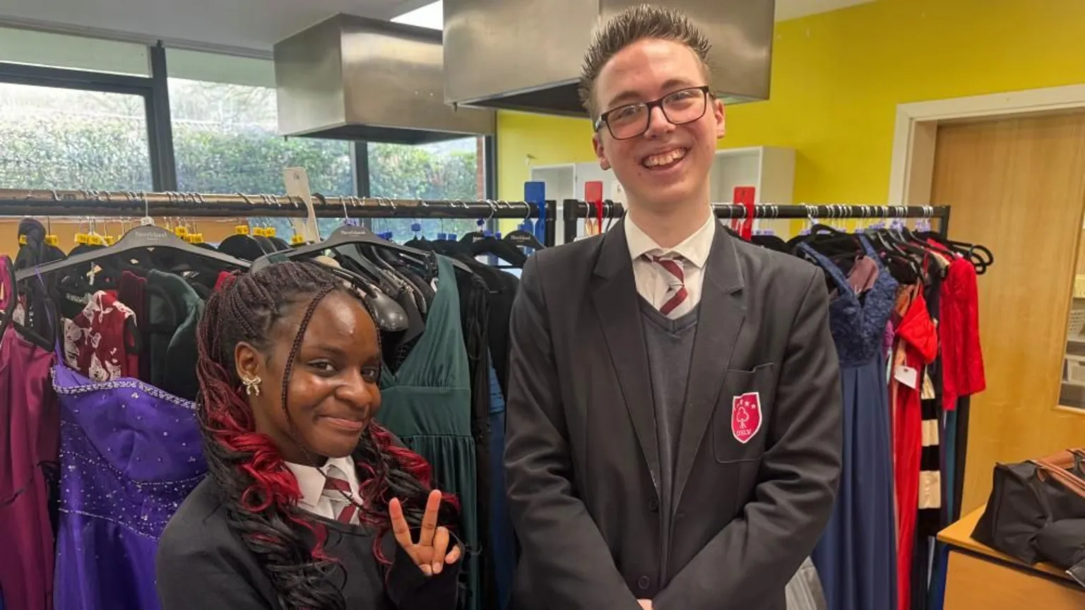 Annabel Amos/BBC Mia and Tate, both in school uniform, in front of a rack of clothes in a room with a wooden door, windows and vents. Mia is wearing a white shirt, tie and jumper, with red in her hair and Tate has short hair, glasses and is wearing a black blazer, white shirt, tie and jumper. 
