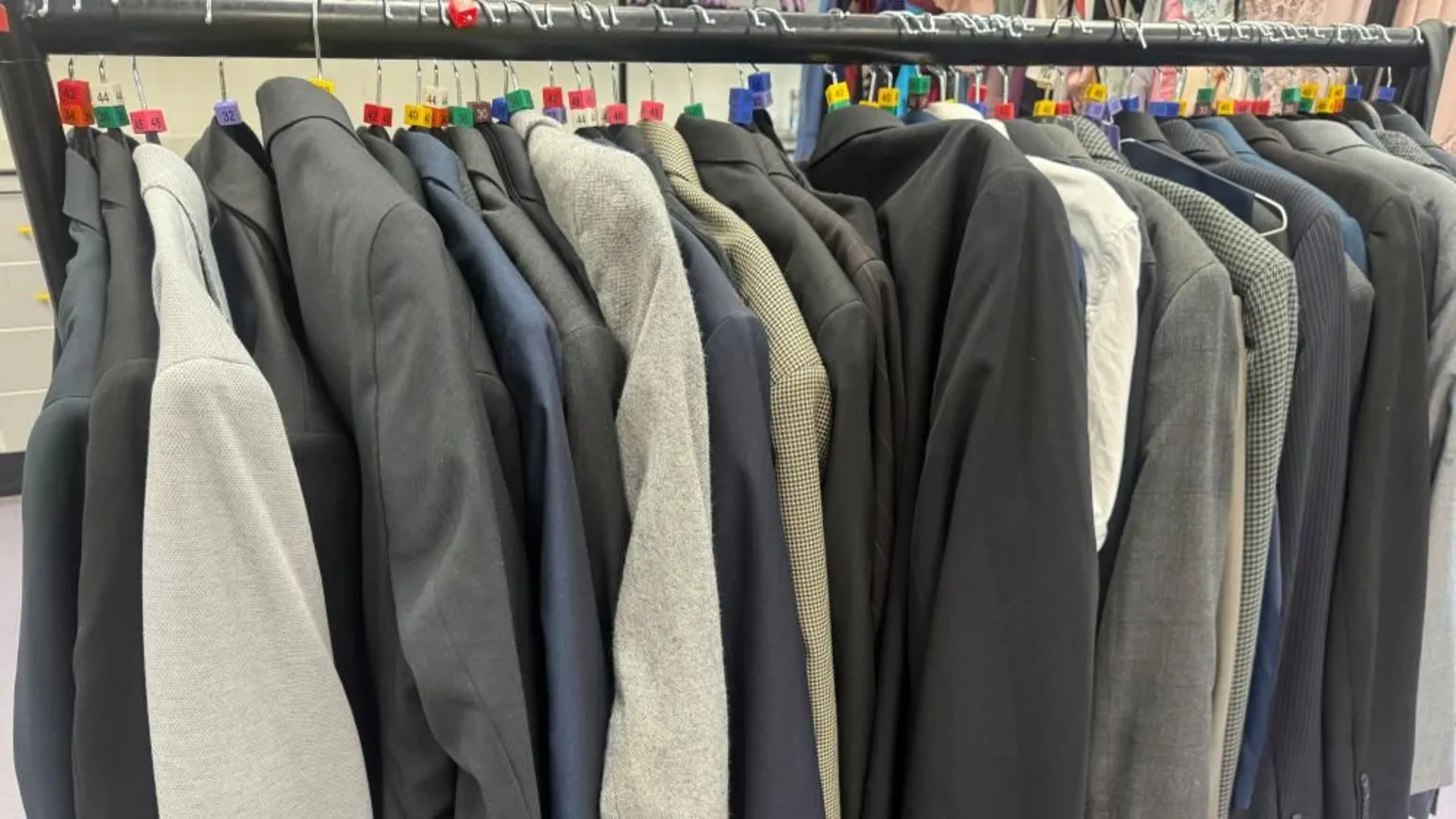 Annabel Amos/BBC A row of suits on a rail in different colours from grey, black and blue. They all have different size number tags on them. 