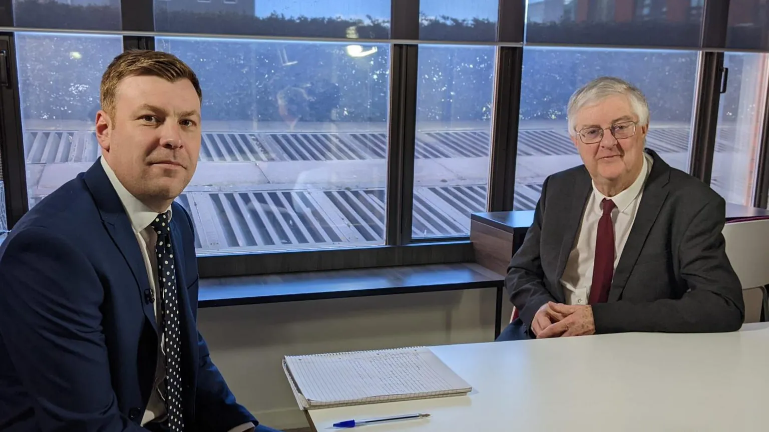 The image shows Mark Drakeford and BBC correspondent Cemlyn Davies sat at a table. Drakeford is wearing a grey suit, white shirt and red tie. Davies is wearing a navy suit, white shirt, and blue and white spotty tie. Both are looking at the camera.