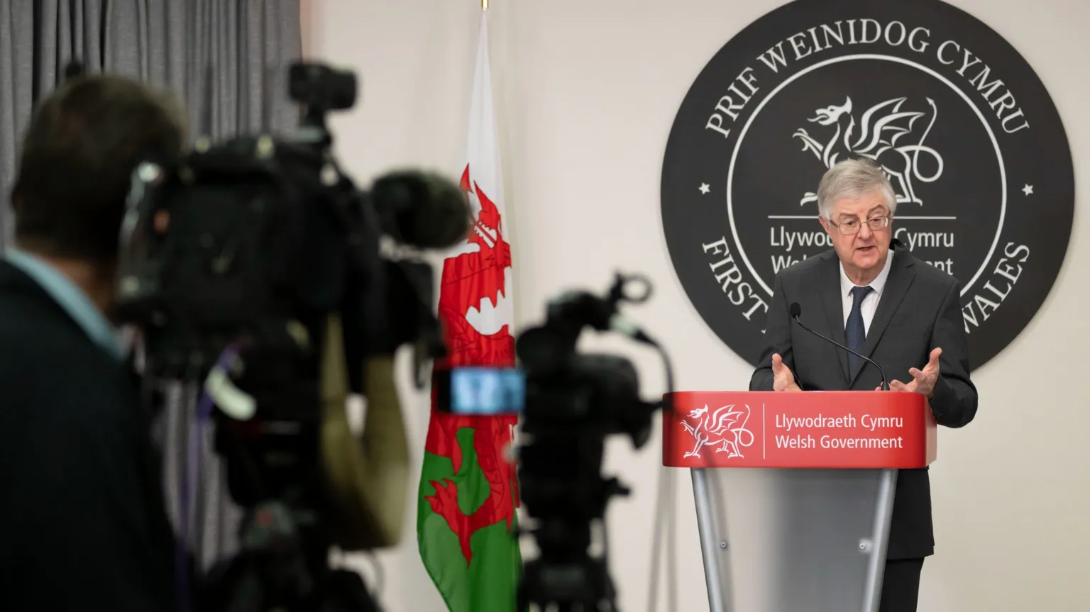 First Minister of Wales Mark Drakeford speaks during a coronavirus press conference, standing in front of a lectern beside a Welsh national flag, and a cameraman operating a camera is seen in the foreground.