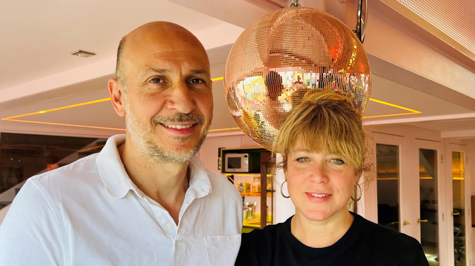 Cobalt Studios Mark Collet standing on the left of Kate Hodgkinson. Collet is bald with grey stubble and is wearing a white shirt. Hodgkinson has light hair with a full fringe which is tied back, hoop earrings and a black top. They are both smiling into the camera. There is a disco ball behind them and some white doors.