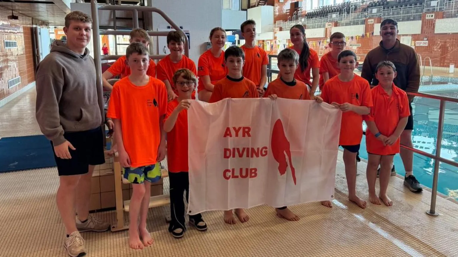 Ayr Diving Club A group of kids wearing orange t-shirts stand by a swimming pool holding a white banner with red block capitals that read - Ayr Diving Club. Two adults stand at either side of the group wearing a dark coloured hoodie and smiling.