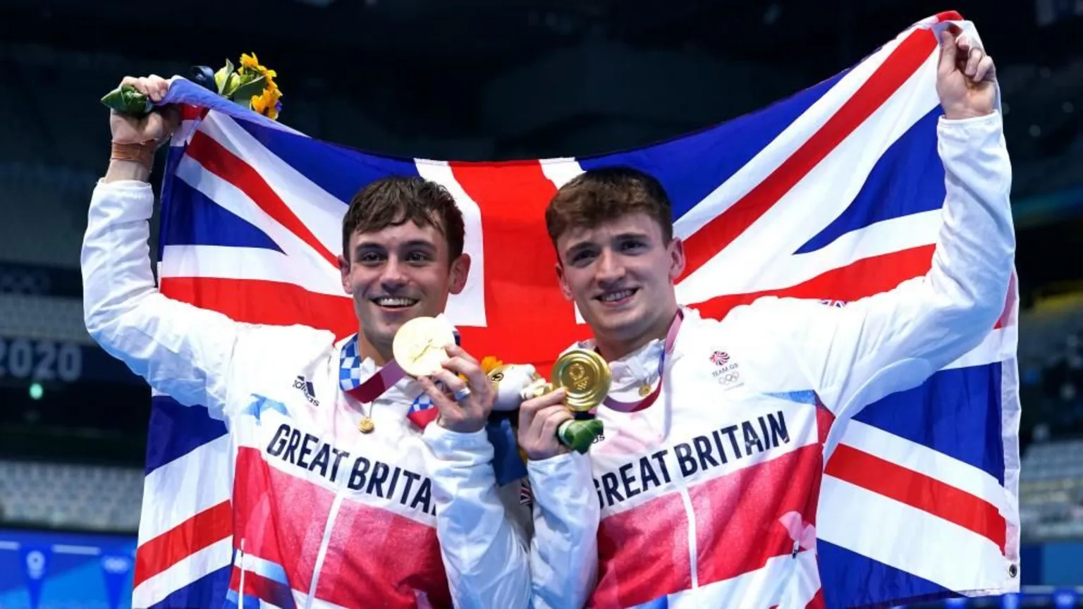 Two young men with brown hair wearing Great Britain track tops holding up a big union jack behind them while holding a gold medal each in their other hands