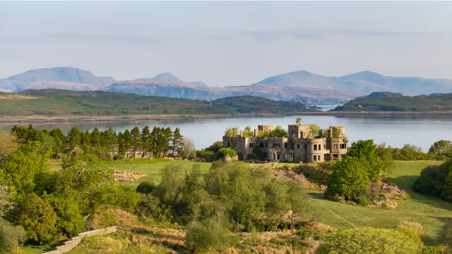Sotheby's/Knight Frank The ruins of Shuna Castle, a flat-roofed turreted castle, are surrounded by grassy banks and trees and overlooking Loch Melfort with a view in the distance of mountains.