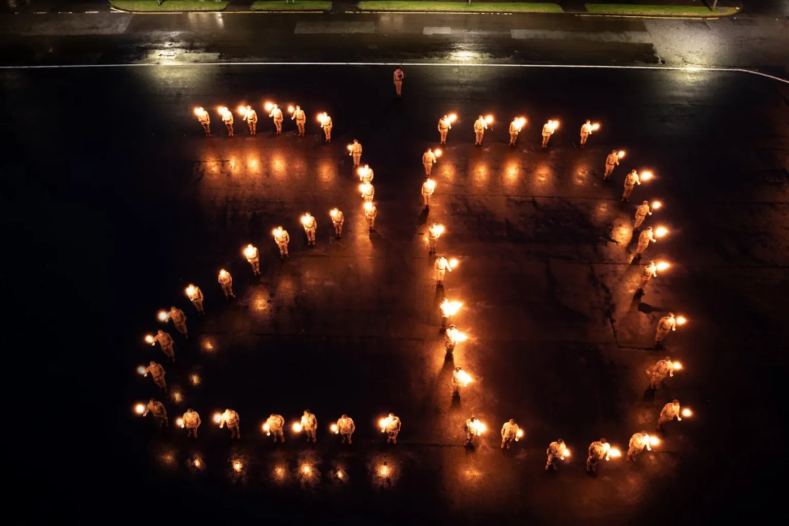  “Large group of uniformed individuals standing outdoors at night, each holding a torch, arranged in formation to create a glowing number ‘20’ on a dark wet surface, with a stone building illuminated in the background.”