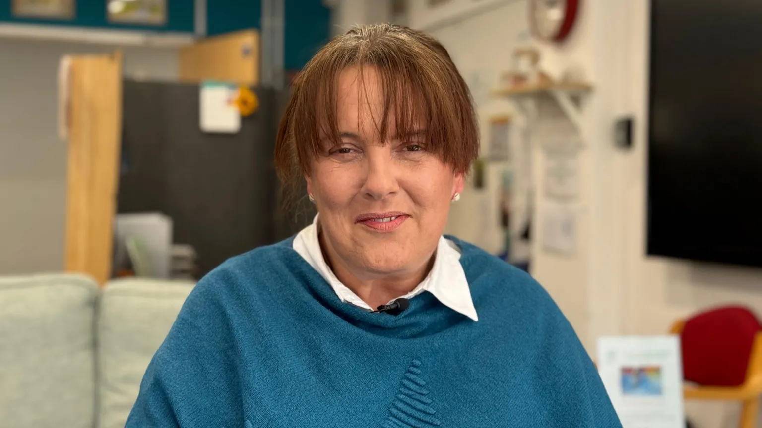 Nicola has a dark brown hair and is smiling as she looks to camera, wearing a blue jumper