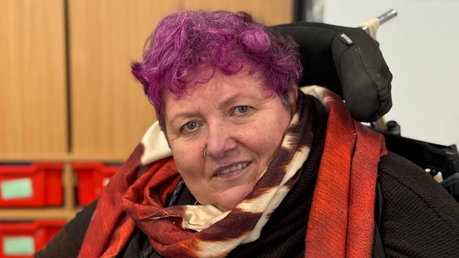 Sarah looks to the camera. She has pink hair and is using a wheelchair. She is wearing a black top and an orange scarf.