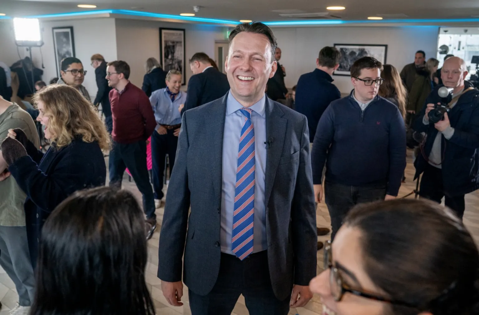  Russell Findlay in a suit and striped tie stands in the middle of a busy indoor event, surrounded by groups of people talking, taking photos and moving around the room, with bright lighting and framed photographs on the walls