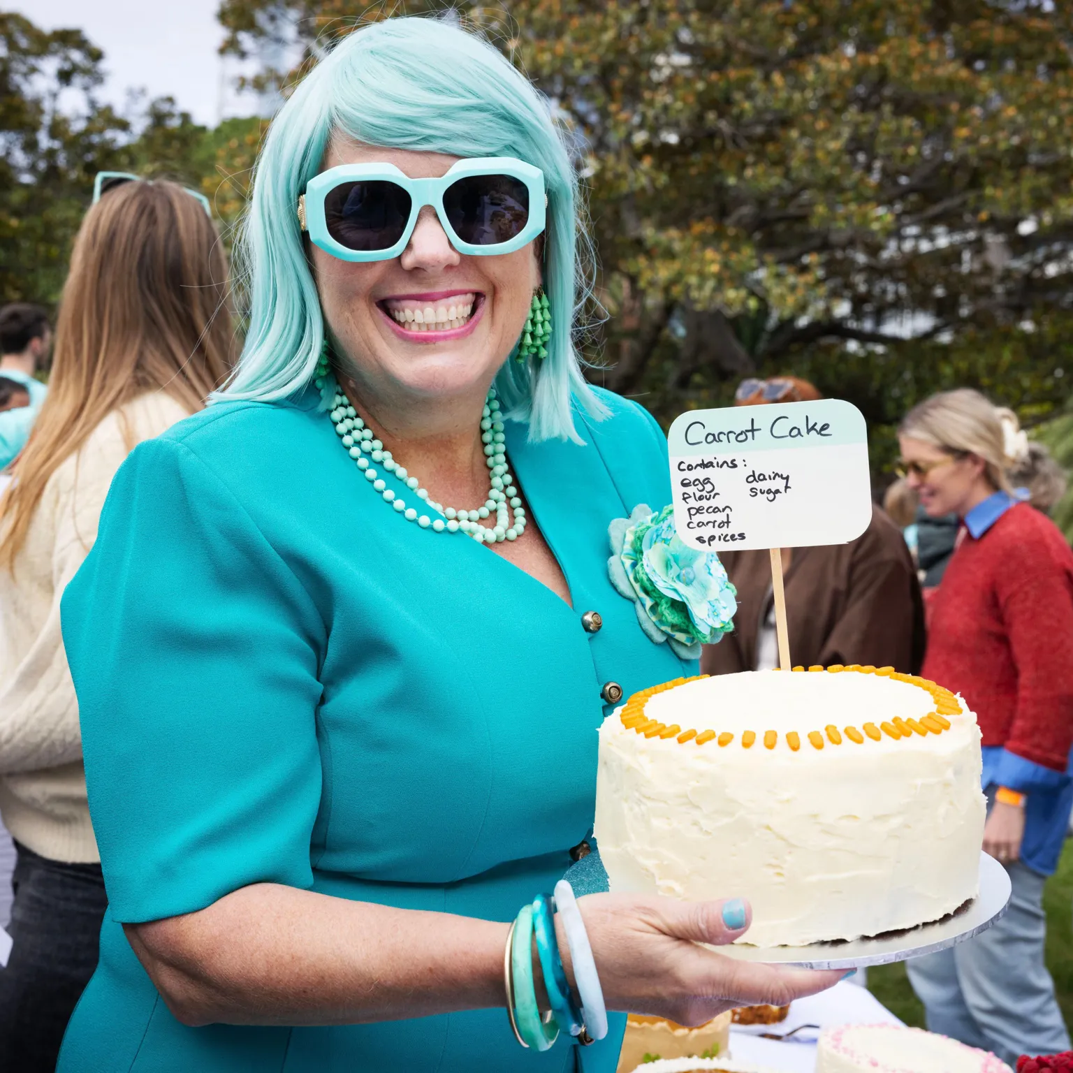Jessica Hromas/BBC A woman with turquoise hair and glasses, green earrings and necklace and a bright turquoise top, with green and turquoise bangles, holding a carrot cake