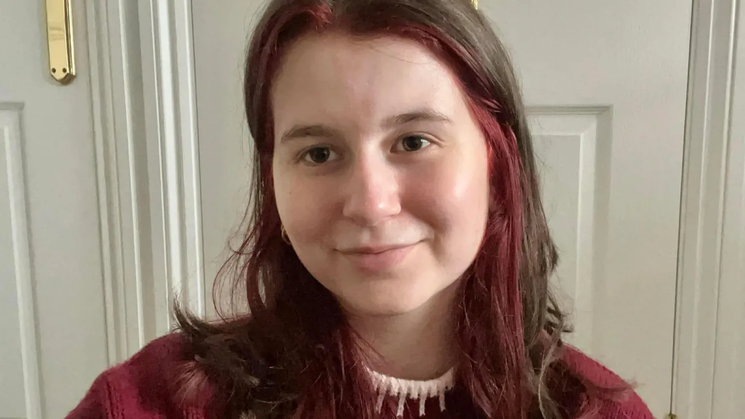 Abi Wilson Selfie photograph of Abi Wilson, 20, who has brown and red hair. She stands in front of a white door frame and smiles at the camera. She has brown eyes and wears a red jumper with a white trim collar.