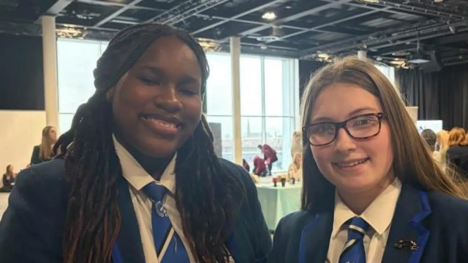 Kayla and Brooke and smiling at the camera. Brooke wears glasses. They are wearing navy school blazers that have lighter blue lining. Their school ties are light blue to match.