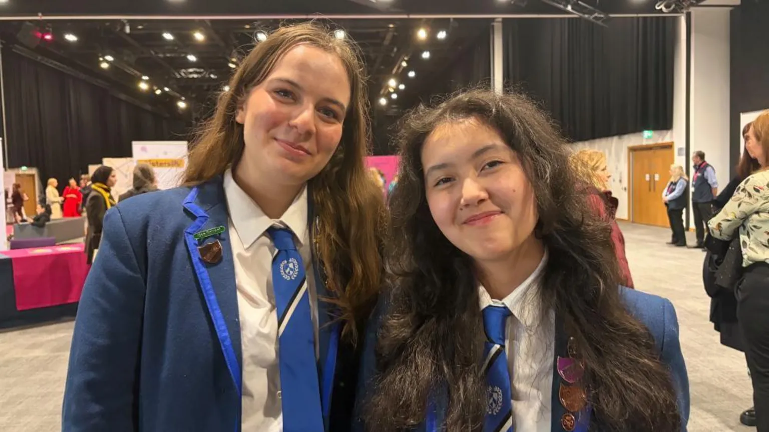 Eve and Anna are smiling at the camera. Eve has light brown hair and Anna has dark brown hair. They are wearing navy school blazers that have lighter blue lining. Their school ties are light blue to match.