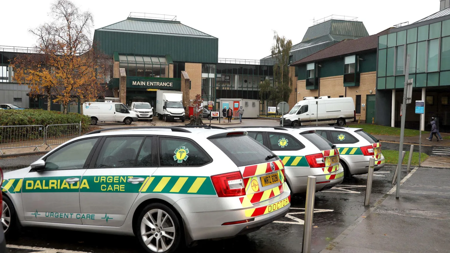 Pacemaker A wide shot of the main entrance to Antrim Area Hospital, a large brown brick building with a green roof. There are several vehicles parked outside the hospital, including three urgent car cars with emergency vehicle markings.