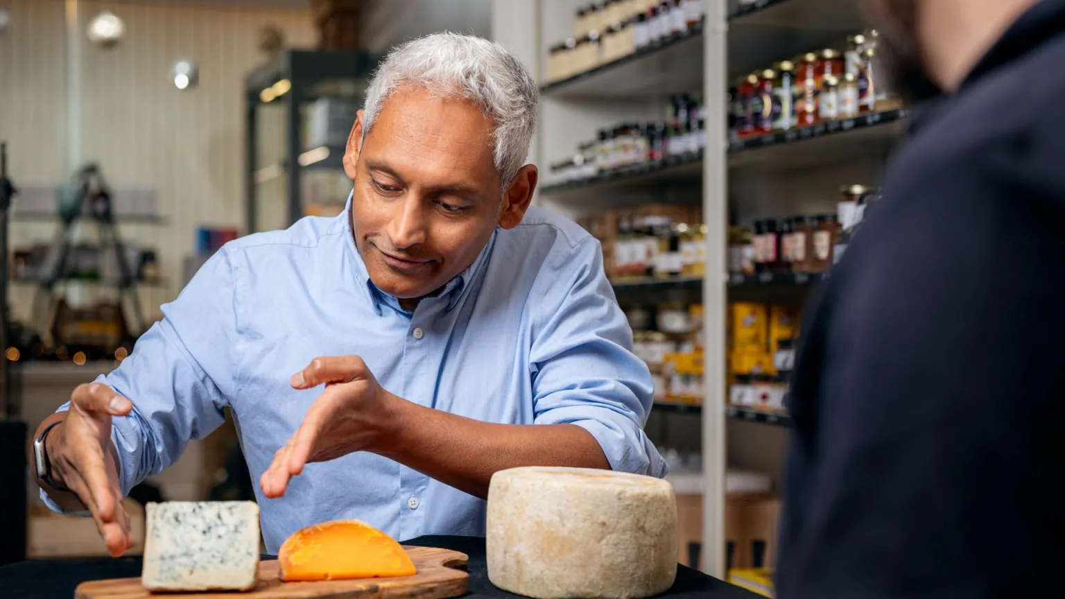 Scott Barron A man with grey hair in a blue shirt points at a block of cheese