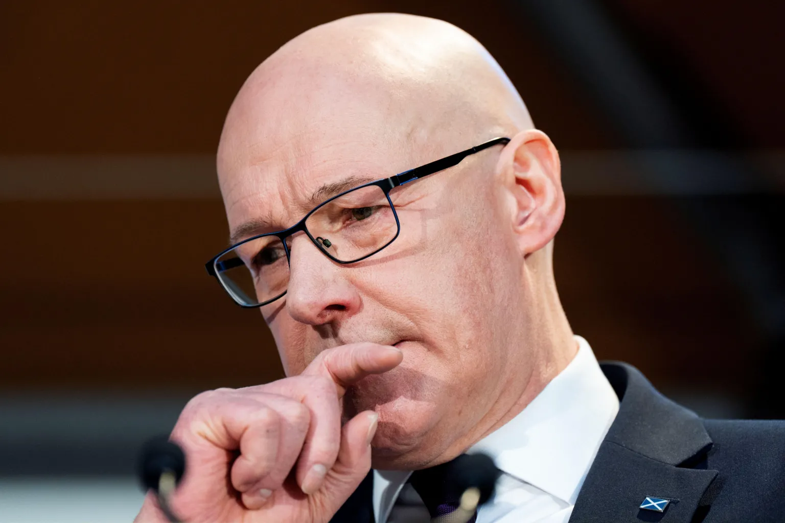 Head shot of John Swinney, who is bald, holds his right hand up to his mouth and looks at the ground with a serious expression on his face.