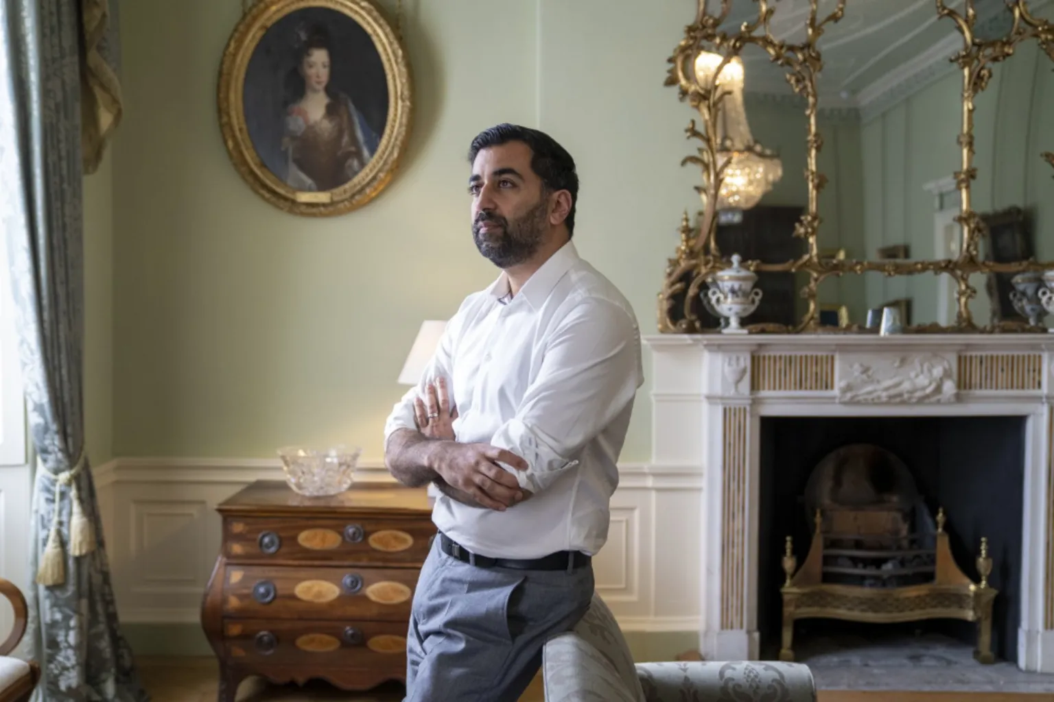  Yousaf standing in an elegant, traditionally decorated room with arms crossed, positioned near a fireplace with ornate gold-framed mirrors and a wooden chest of drawers beneath a portrait on the wall.