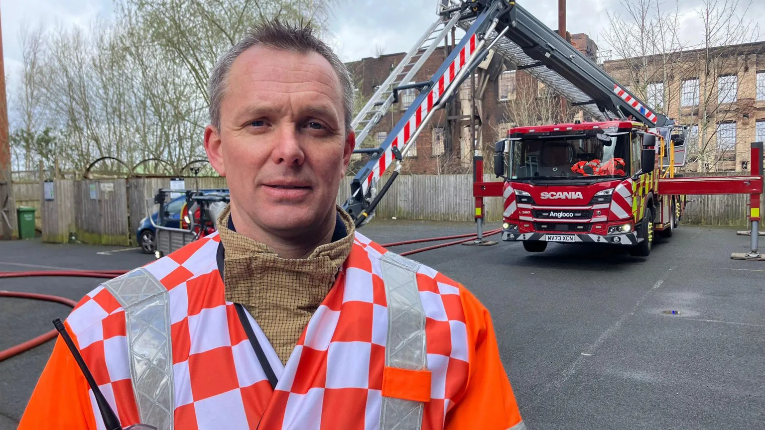 A man in an orange and white check jacket with high-vis stripes either side of the collar. He stands in front of a fire engine with ladder extended. A large mill building can be seen behind.