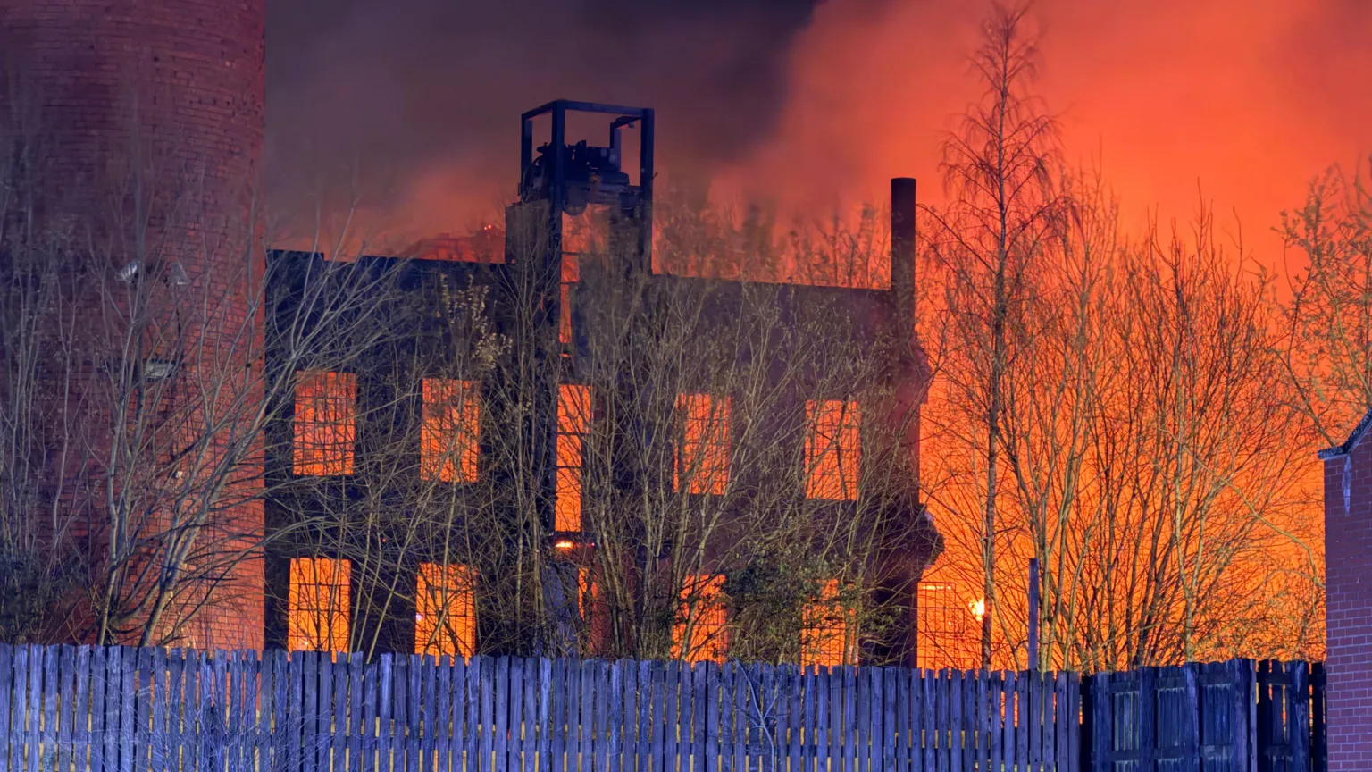 A large blaze at night in a mill building. The sky is lit up orange. Wooden fencing is in front of the building.