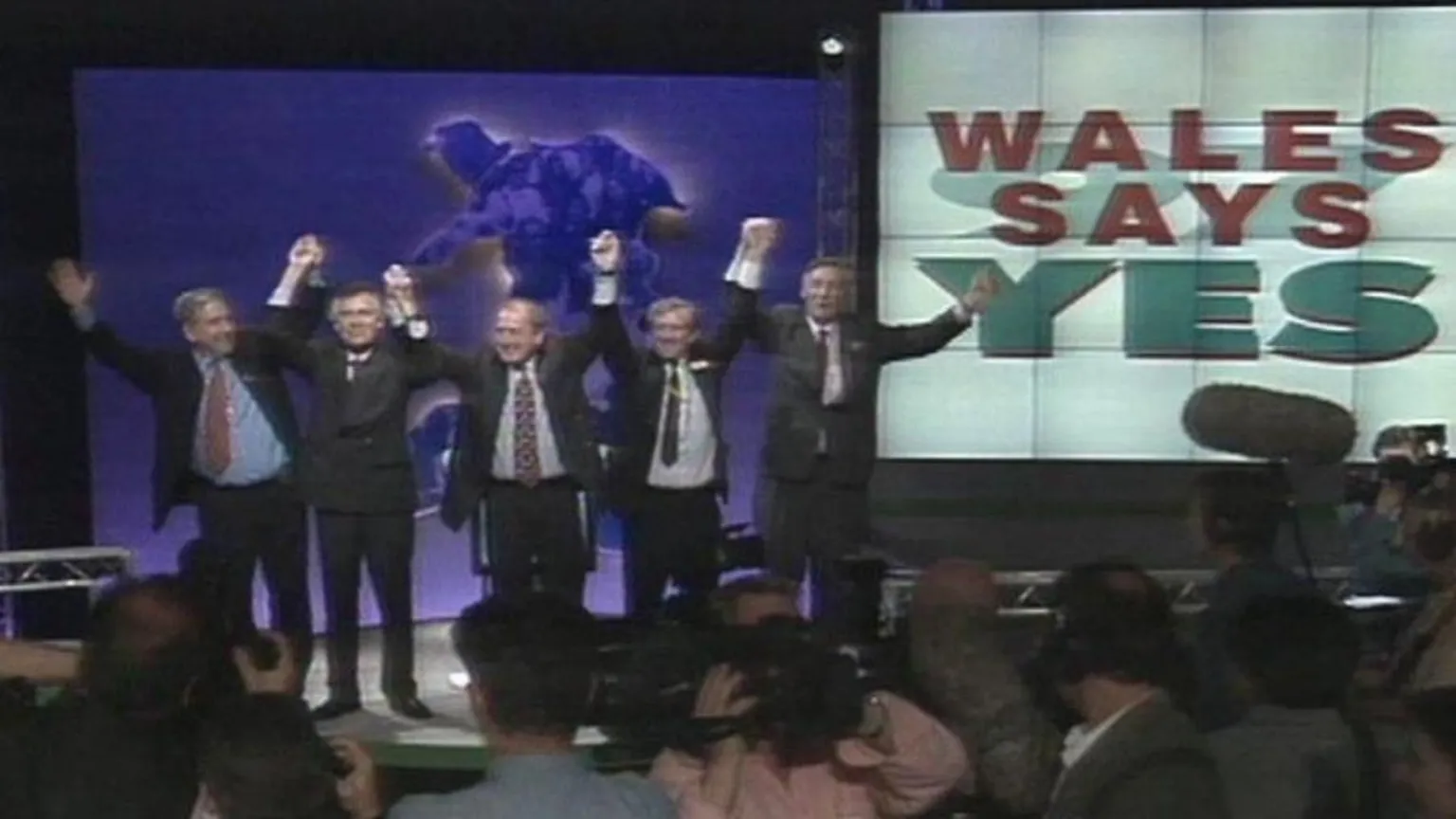 Four prominent figures from the Yes campaign stand on a stage in front of a crows of television cameras. The have their arms aloft and are smiling. Behind them is a screen showing the outline of Wales. On a separate screen are the words: 'Wales says yes'.