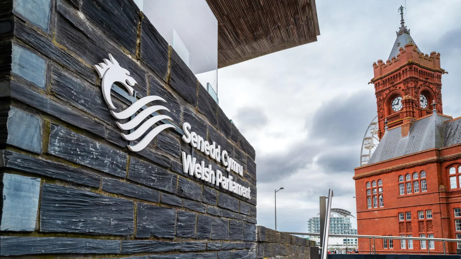  Part of the Senedd building in Cardiff Bay is prominent to the left and in the foreground of the image. The Senedd's logo can be seen on the building slate wall along with the institution's name in both languages. The right of the image is taken up by the red brick Pierhead building. It is a cloudy day.