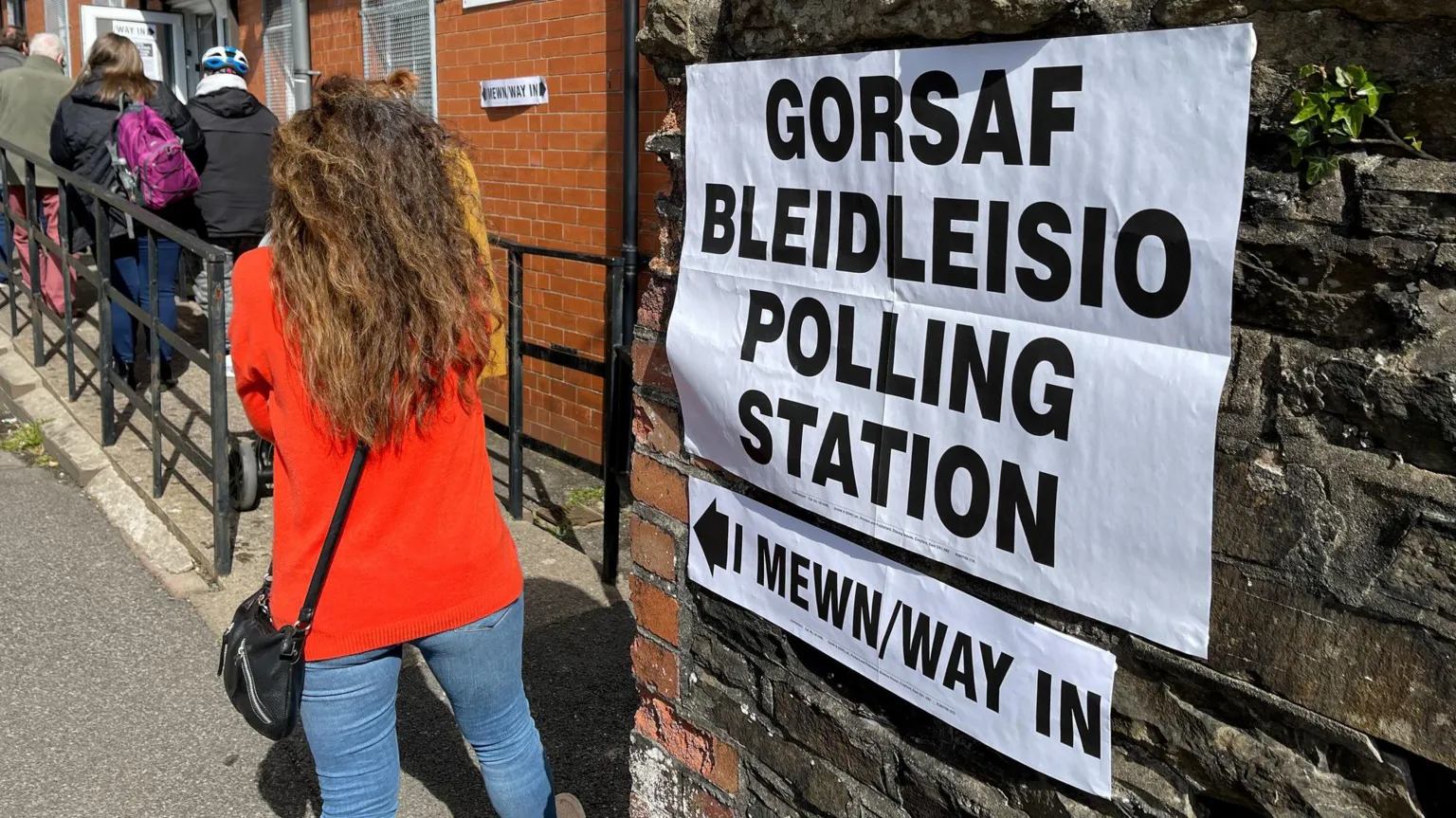  The image shows voters waiting to enter a polling station. There is a bilingual notice on a stone wall in the foreground letting people know that they are at a polling station and how they should enter. It is a sunny day.